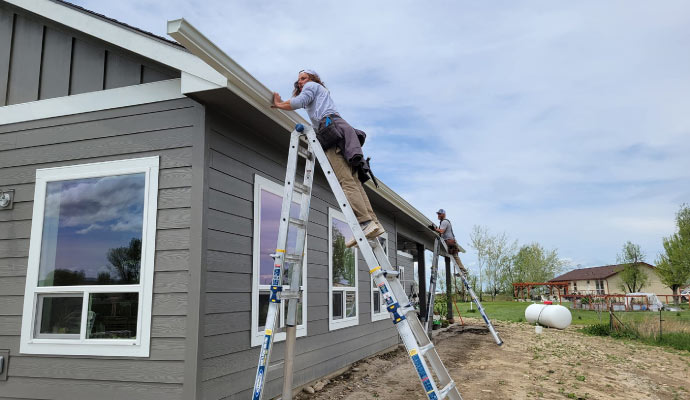 Two individuals on ladders installing a gutter on the edge of house's roof.