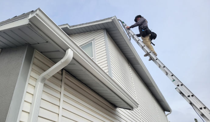 A person on ladder inspecting a gutter on the house roof.